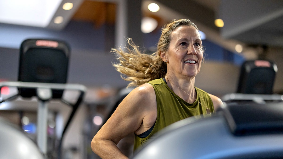 A woman working out indoors on a treadmill, she is wearing a green tank top and smiling - we see her from the chest up.