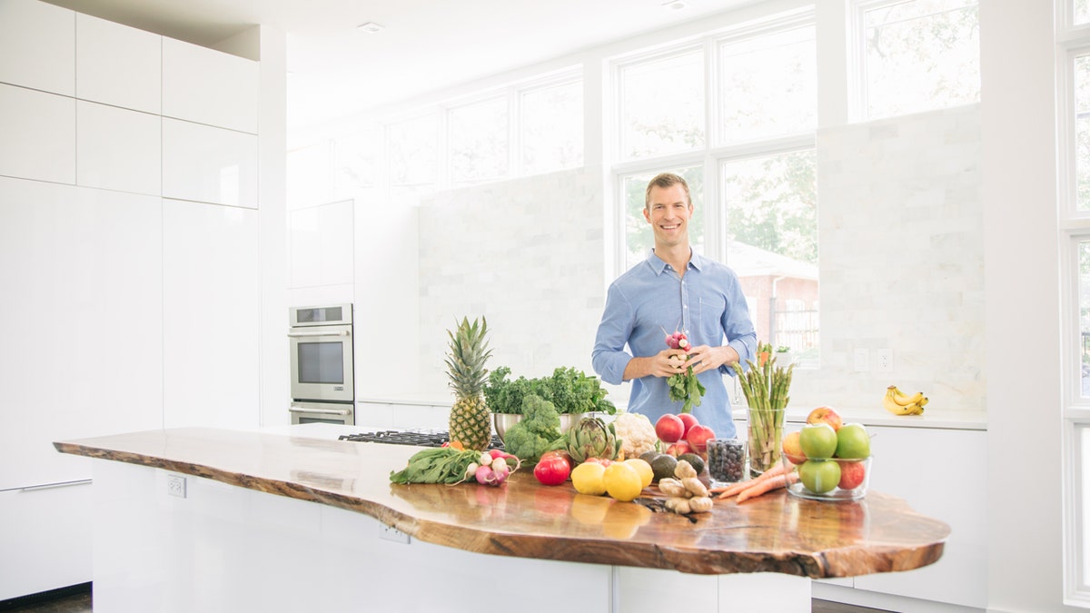Dr. Axe standing in a white kitchen with a table covered in fruits and vegetables