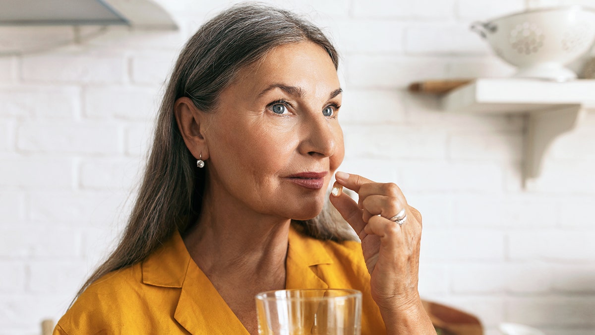 Older woman holding a capsule pill near her mouth with a glass of water in hand.