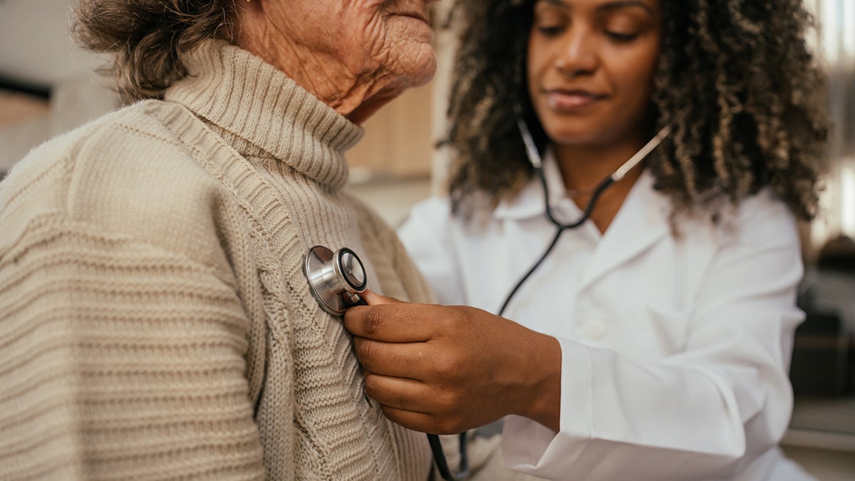 Young female doctor listens to older woman's heart at appointment using stethoscope.