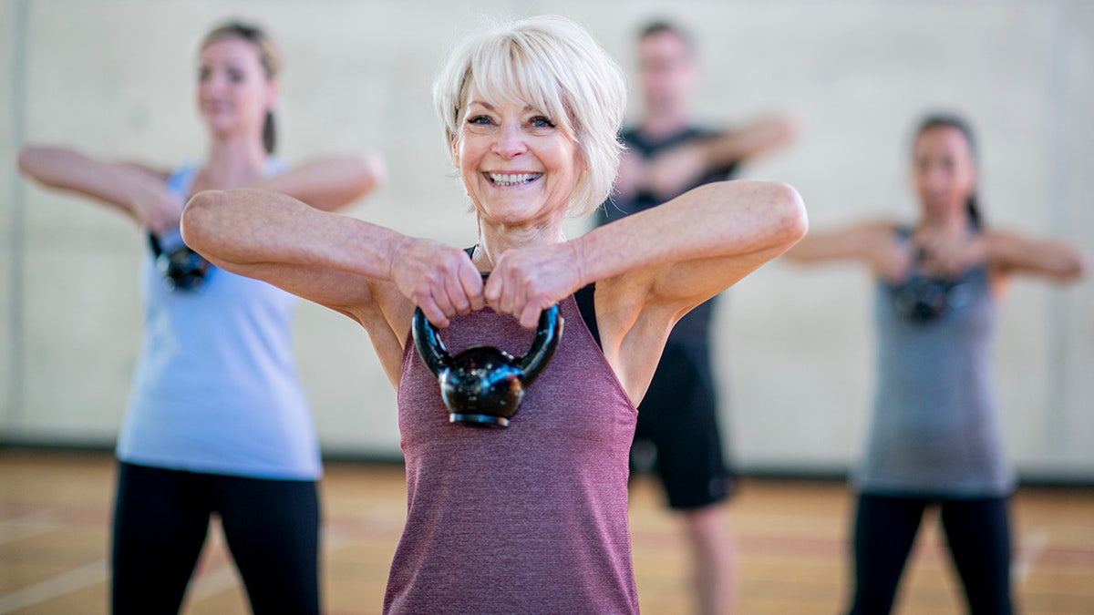Older woman seen smiling with kettle bell held up to her chest in workout class.