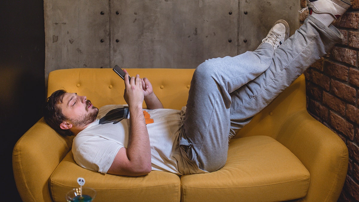 Young man lying on the yellow sofa, spending his leisure time resting, looking at phone with feet up against wall.