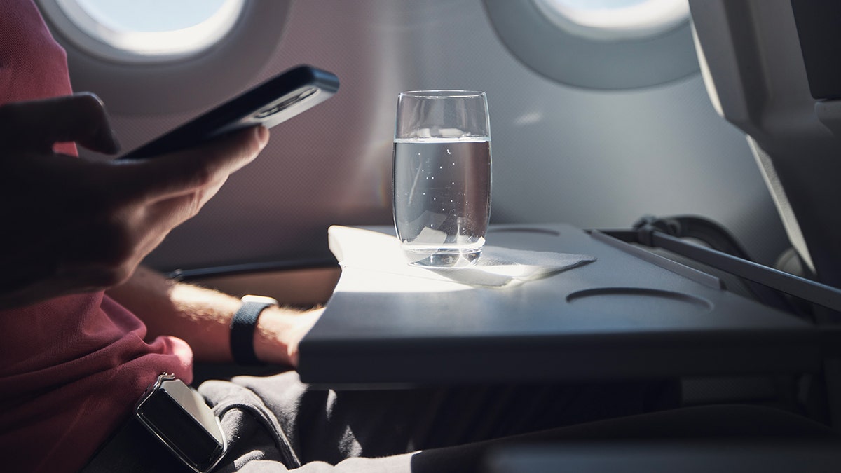 Passenger using a smartphone next to a glass of water on an airplane tray table by the window.