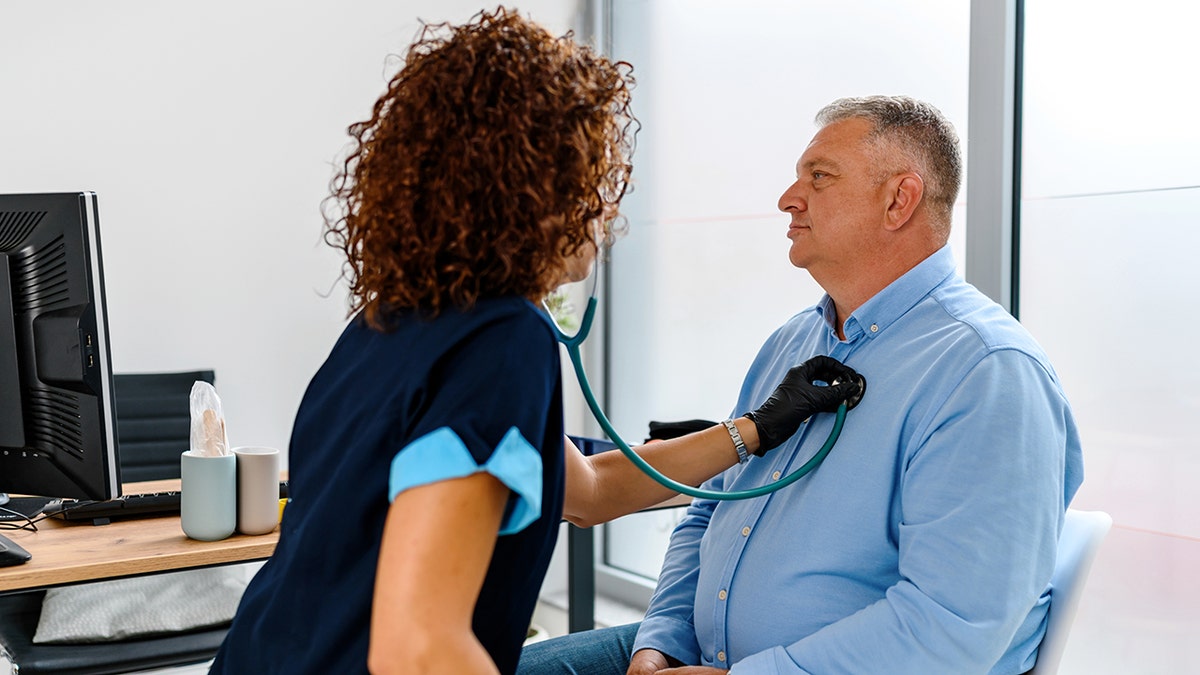 Nurse using a stethoscope to examine an adult male patient during a medical checkup in a clinic exam room.