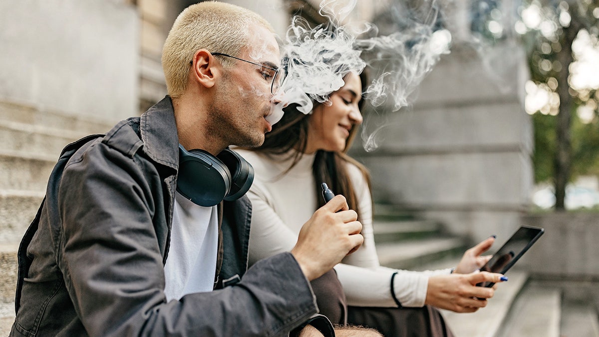 Two college students sitting outside on stairs as man vapes and woman looks at digital tablet.