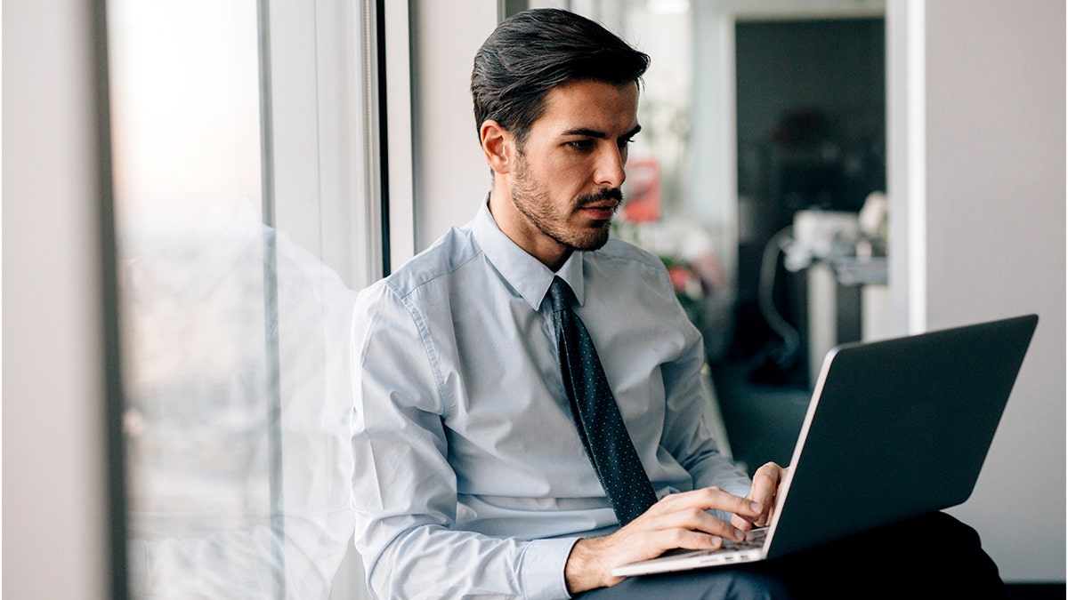 A man sits in front of a window, working on a laptop.