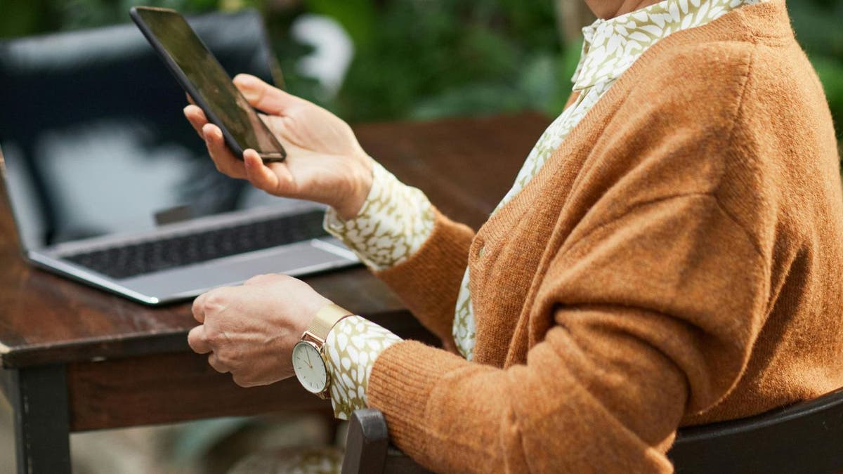 A woman holds a phone in front of a laptop.