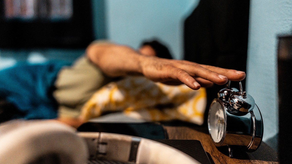 Man's hand seen hitting alarm clock from bed.