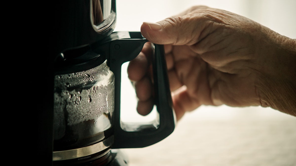 Older man's hand grabbing coffee carafe handle up-close.